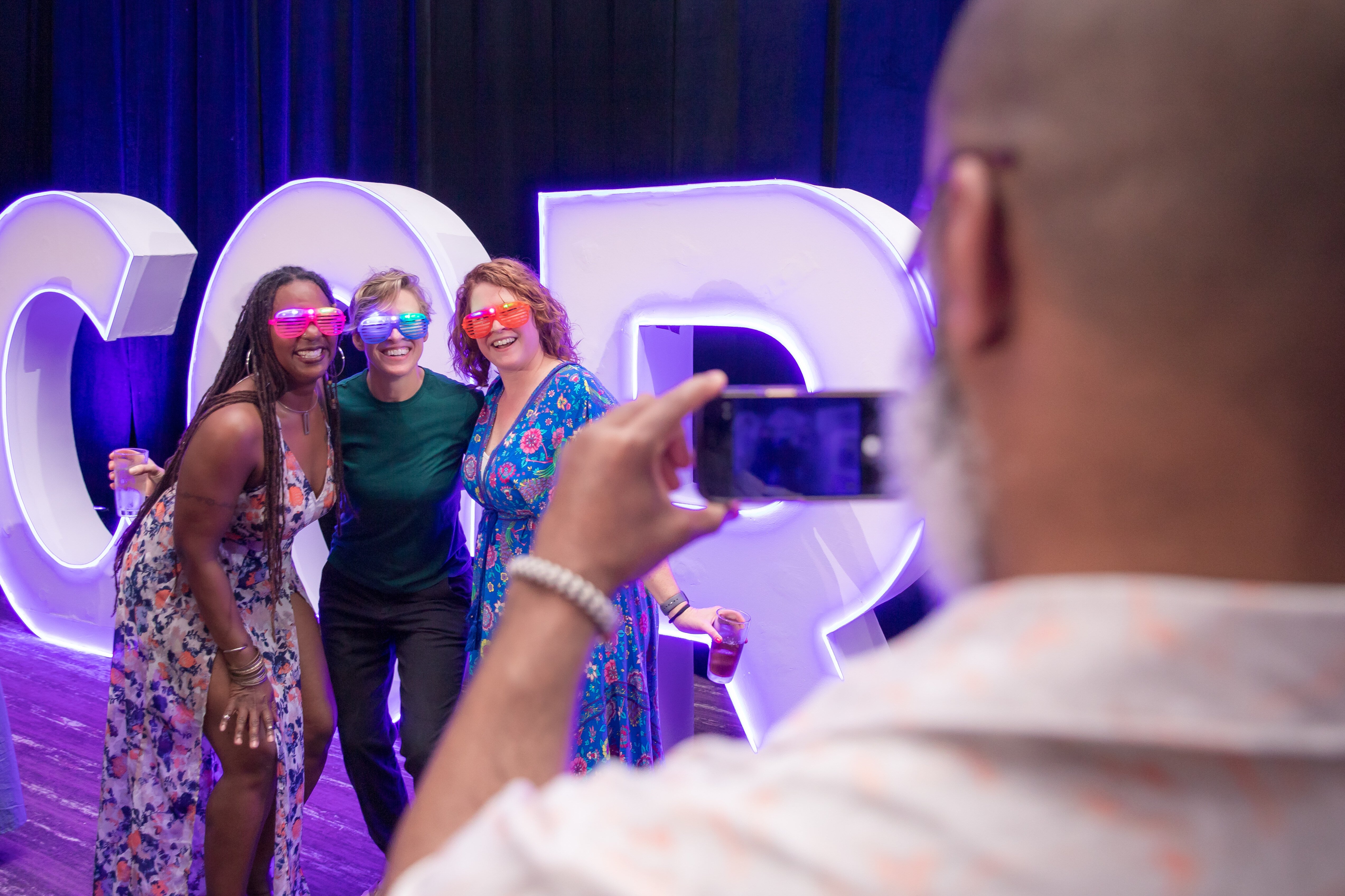 Person taking a photo of three individuals with fun sunglasses on.